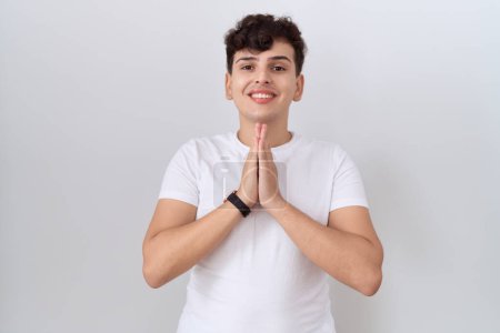 Foto de Joven hombre no binario vistiendo casual camiseta blanca rezando con las manos juntas pidiendo perdón sonriendo confiado. - Imagen libre de derechos