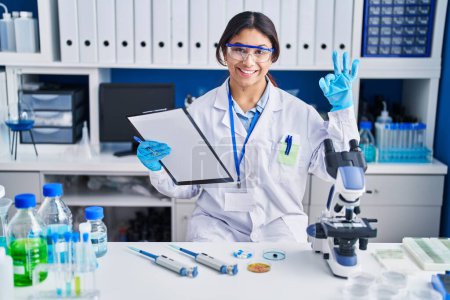 Foto de Mujer joven hispana que trabaja en el laboratorio científico sonriendo positiva haciendo signo de ok con la mano y los dedos. expresión exitosa. - Imagen libre de derechos