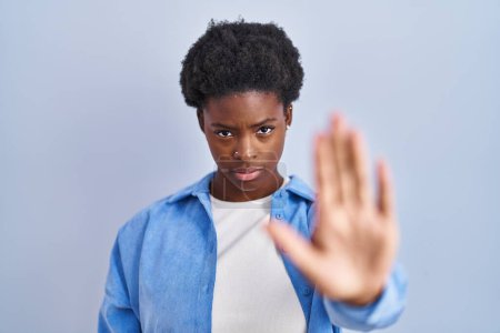 Téléchargez les photos : Femme afro-américaine debout sur fond bleu faire arrêter chanter avec la paume de la main. expression d'avertissement avec geste négatif et sérieux sur le visage. - en image libre de droit
