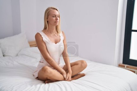 Téléchargez les photos : Jeune femme blonde faisant de l'exercice de yoga assis sur le lit dans la chambre - en image libre de droit