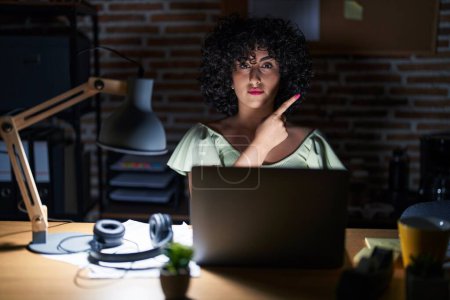 Téléchargez les photos : Jeune femme brune aux cheveux bouclés travaillant au bureau la nuit pointant du doigt vers le côté montrant de la publicité, visage sérieux et calme - en image libre de droit