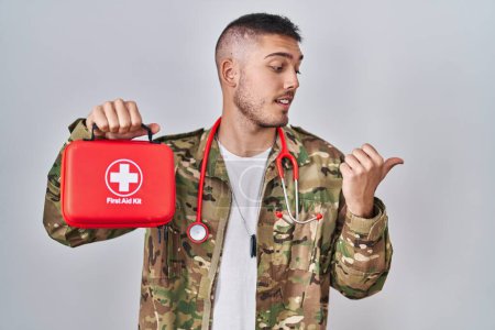 Photo for Young hispanic doctor wearing camouflage army uniform holding first aid kit pointing thumb up to the side smiling happy with open mouth - Royalty Free Image