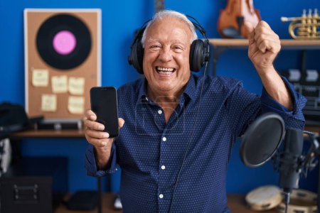 Photo for Senior man with grey hair showing smartphone screen at music studio celebrating victory with happy smile and winner expression with raised hands - Royalty Free Image