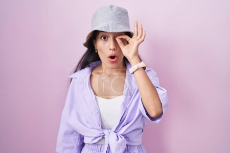 Photo for Young hispanic woman standing over pink background wearing hat doing ok gesture shocked with surprised face, eye looking through fingers. unbelieving expression. - Royalty Free Image