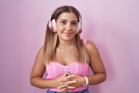 Foto de Mujer rubia joven escuchando música usando auriculares con las manos juntas y los dedos cruzados sonriendo relajada y alegre. éxito y optimismo - Imagen libre de derechos