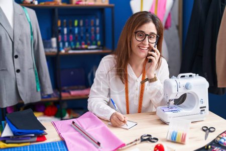 Photo for Young woman tailor talking on smartphone writing on notebook at sewing studio - Royalty Free Image