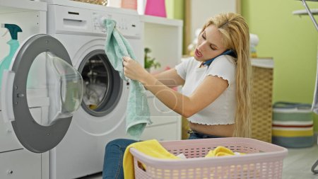 Téléchargez les photos : Jeune femme blonde parlant sur smartphone laver les vêtements à la buanderie - en image libre de droit