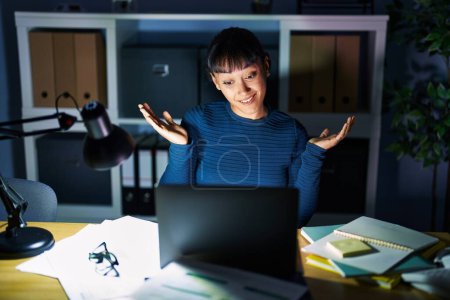Photo for Young beautiful woman working at the office at night clueless and confused expression with arms and hands raised. doubt concept. - Royalty Free Image