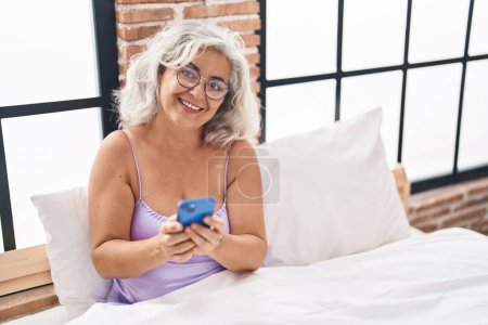 Foto de Mujer de pelo gris de mediana edad con teléfono inteligente sentado en la cama en el dormitorio - Imagen libre de derechos