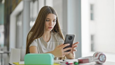 Téléchargez les photos : Jeune belle étudiante utilisant smartphone regardant bouleversé à la bibliothèque - en image libre de droit