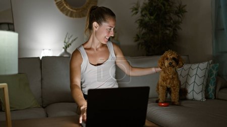 Téléchargez les photos : Jeune femme caucasienne avec chien souriant à l'aide d'un ordinateur portable assis sur le canapé à la maison - en image libre de droit