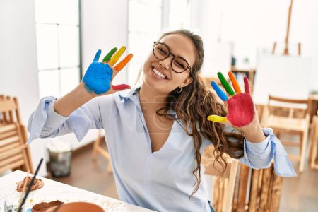 Téléchargez les photos : Jeune belle artiste hispanique femme souriante confiante montrant les mains peintes au studio d'art - en image libre de droit