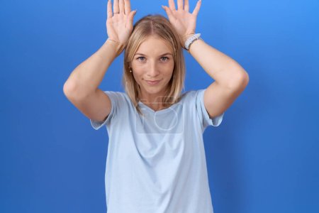 Téléchargez les photos : Jeune femme caucasienne portant un t-shirt bleu décontracté faisant geste d'oreilles de lapin avec les paumes des mains regardant cynique et sceptique. concept lapin de Pâques. - en image libre de droit