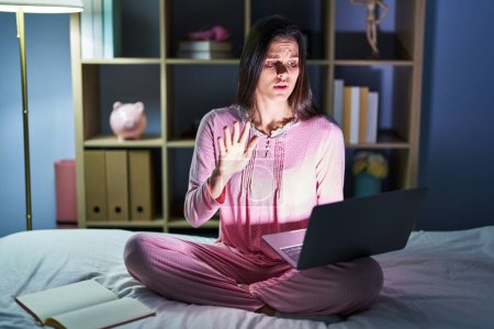 Photo for Young hispanic woman using computer laptop on the bed doing stop gesture with hands palms, angry and frustration expression - Royalty Free Image