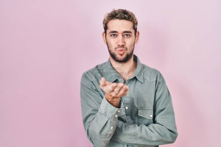 Foto de Hombre hispano con barba de pie sobre fondo rosa mirando a la cámara soplando un beso con la mano en el aire siendo encantador y sexy. expresión de amor. - Imagen libre de derechos