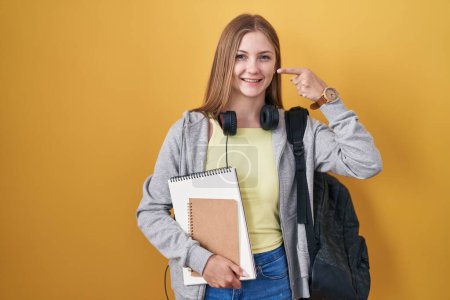 Foto de Young caucasian woman wearing student backpack and holding books pointing with hand finger to face and nose, smiling cheerful. beauty concept - Imagen libre de derechos