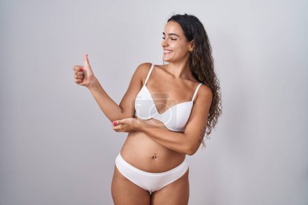 Foto de Mujer hispana joven vistiendo lencería blanca que se ve orgullosa, sonriendo haciendo un gesto hacia un lado - Imagen libre de derechos