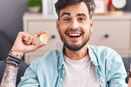 Foto de Joven hombre hispano con tatuajes sosteniendo la moneda criptomoneda que celebra el logro con sonrisa feliz y expresión ganadora con la mano levantada - Imagen libre de derechos