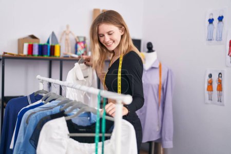 Téléchargez les photos : Jeune femme blonde tailleur souriant confiant tenant des vêtements sur rack au magasin de tailleur - en image libre de droit