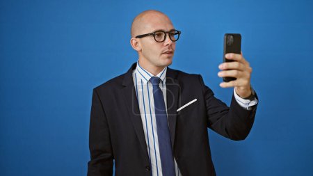 Foto de Hombre hispano joven trabajador de negocios con cara seria hacer selfie por teléfono inteligente sobre fondo azul aislado - Imagen libre de derechos