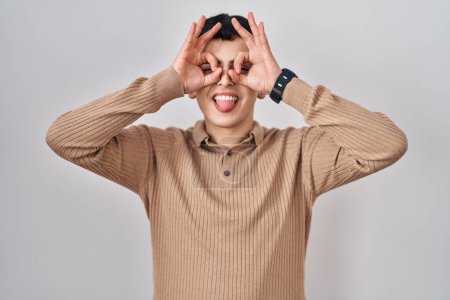 Photo for Non binary person standing over isolated background doing ok gesture like binoculars sticking tongue out, eyes looking through fingers. crazy expression. - Royalty Free Image