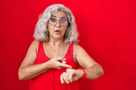 Foto de Mujer de mediana edad con el pelo gris de pie sobre el fondo rojo en la prisa que apunta a ver el tiempo, la impaciencia, molesto y enojado por retraso plazo - Imagen libre de derechos