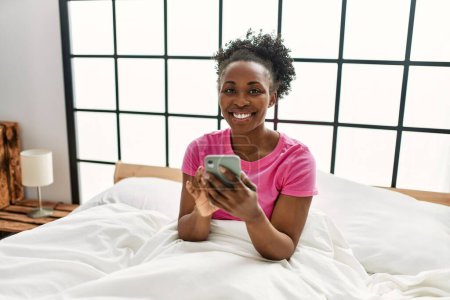 Téléchargez les photos : Femme afro-américaine utilisant smartphone assis sur le lit dans la chambre - en image libre de droit