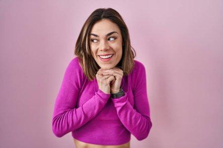 Téléchargez les photos : Femme hispanique debout sur fond rose riant nerveux et excité avec les mains sur le menton regardant vers le côté - en image libre de droit
