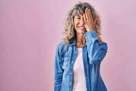 Téléchargez les photos : Femme d'âge moyen debout sur fond rose couvrant un oeil avec la main, sourire confiant sur le visage et l'émotion surprise. - en image libre de droit
