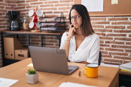 Photo for Hispanic young woman working at the office wearing glasses thinking concentrated about doubt with finger on chin and looking up wondering - Royalty Free Image