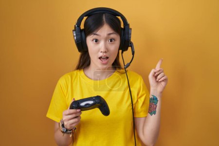 Téléchargez les photos : Chinoise jeune femme jouant jeu vidéo tenant contrôleur surpris pointant du doigt sur le côté, bouche ouverte expression étonnante. - en image libre de droit