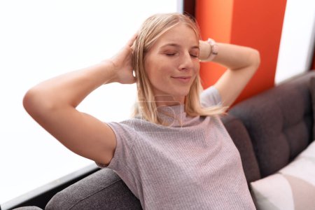 Téléchargez les photos : Jeune femme blonde détendue avec les mains sur la tête assise sur le canapé à la maison - en image libre de droit