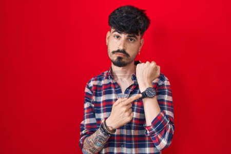 Téléchargez les photos : Jeune homme hispanique avec la barbe debout sur fond rouge à la hâte pointant vers regarder le temps, l'impatience, regardant la caméra avec une expression détendue - en image libre de droit