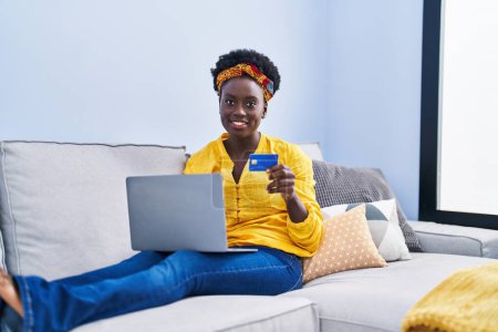 Téléchargez les photos : Jeune femme afro-américaine utilisant ordinateur portable et carte de crédit assis sur le canapé à la maison - en image libre de droit
