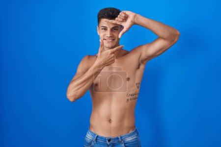 Téléchargez les photos : Jeune homme hispanique debout torse nu sur fond bleu souriant faisant cadre avec les mains et les doigts avec le visage heureux. concept de créativité et photographie. - en image libre de droit