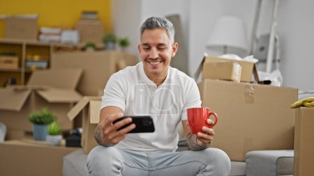 Photo for Young hispanic man drinking coffee watching video on smartphone at new home - Royalty Free Image