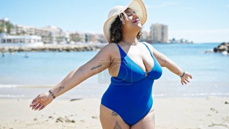 Foto de Joven hermosa turista latina con traje de baño y sombrero de verano de pie con los brazos abiertos en la playa - Imagen libre de derechos