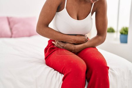 Téléchargez les photos : Femme afro-américaine souffrant de douleurs menstruelles assise sur le lit dans la chambre - en image libre de droit