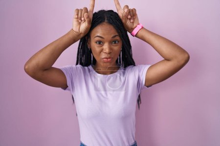 Téléchargez les photos : Femme afro-américaine avec des tresses debout sur fond rose faisant un geste drôle avec le doigt sur la tête comme des cornes de taureau - en image libre de droit