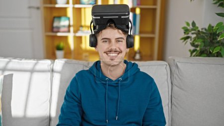 Téléchargez les photos : Jeune homme hispanique portant des lunettes de réalité virtuelle souriant à la maison - en image libre de droit
