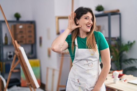 Téléchargez les photos : Jeune femme brune au studio d'art souriant avec la main sur l'oreille écoutant et entendant des rumeurs ou des commérages. concept de surdité. - en image libre de droit