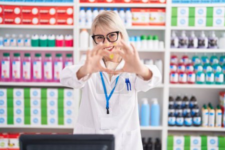 Téléchargez les photos : Young caucasian woman working at pharmacy drugstore smiling in love doing heart symbol shape with hands. romantic concept. - en image libre de droit