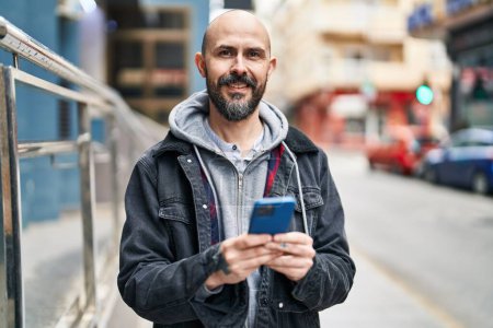 Téléchargez les photos : Jeune homme chauve souriant confiant en utilisant un smartphone dans la rue - en image libre de droit
