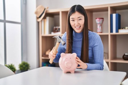 Photo for Chinese young woman holding hammer and piggy bank smiling and laughing hard out loud because funny crazy joke. - Royalty Free Image