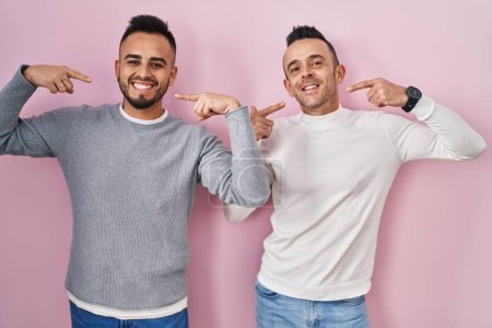 Téléchargez les photos : Couple homosexuel debout sur fond rose souriant gai montrant et pointant avec les doigts dents et la bouche. concept de santé dentaire. - en image libre de droit