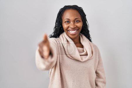Foto de Mujer africana de pie sobre fondo blanco sonriendo amistoso ofreciendo apretón de manos como saludo y bienvenida. negocios exitosos. - Imagen libre de derechos