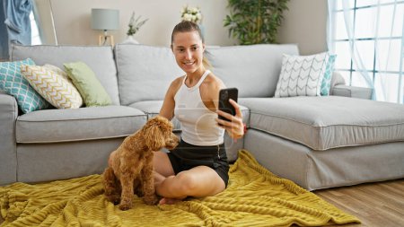 Foto de Joven mujer caucásica con perro sonriendo tomando foto selfie con teléfono inteligente sentado en el suelo en casa - Imagen libre de derechos