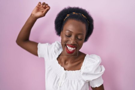Téléchargez les photos : Femme africaine aux cheveux bouclés debout sur fond rose dansant heureux et joyeux, souriant décontracté et confiant écoutant de la musique - en image libre de droit