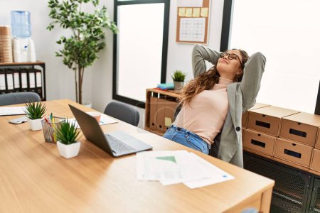 Foto de Joven hermosa mujer hispana trabajadora de negocios relajada con las manos en la cabeza en la oficina - Imagen libre de derechos