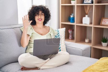 Téléchargez les photos : Jeune femme brune aux cheveux bouclés utilisant un ordinateur portable assis sur le canapé à la maison montrant et pointant vers le haut avec les doigts numéro cinq tout en souriant confiant et heureux. - en image libre de droit